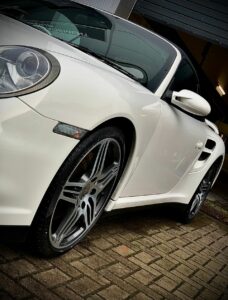 Close-up shot of a white sports car parked on a brick driveway. The image focuses on the sleek design, front right headlight, and the intricate details of the alloy wheels.