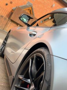 A close-up view of a sleek, modern sports car with a matte gray finish. The image focuses on the rear wheel and side detailing, showcasing the car’s aerodynamic curves and black Pirelli tires. A brick building with a peaked roof is visible in the background.