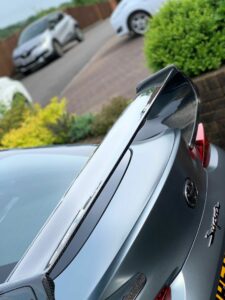 Close-up of the rear of a gray sports car, focusing on the carbon fiber spoiler and the "Supra" emblem. In the background, a curved driveway is lined with bushes and other parked cars. The setting appears to be a residential area.