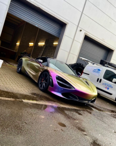 A sleek, iridescent sports car with a gradient paint job from purple to gold is parked in front of a gray industrial building. The car is low to the ground with an aerodynamic design. Next to it, a white van is parked, partially visible. The ground appears wet.