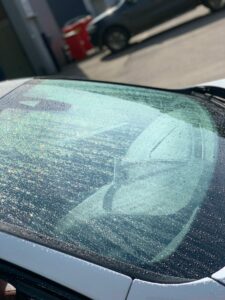 A close-up shot of a car windshield covered in water droplets, with the sunlight causing a sparkling effect. The car hood and part of another vehicle are visible in the background under a clear sky.