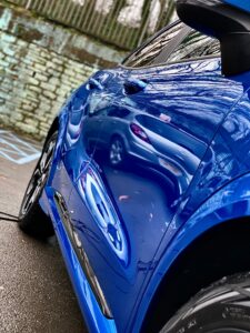 A close-up shot of a blue car with reflections of the surrounding environment on its shiny surface. The background features a stone wall with a fence on top and some trees, and the ground is wet, indicating recent rain.