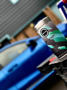 Close-up of a white and turquoise container with the label "Synergy 2 Super Ceramic Coating" on a black surface. In the background, there's a slightly blurred blue car parked beside a building. The image focuses on the container while the car is out of focus.