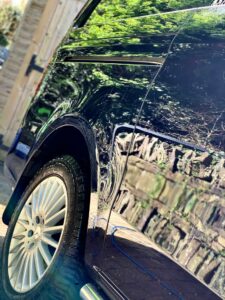 Close-up of a sleek, dark-colored car showcasing the front wheel and part of the side body. The car's shiny surface reflects its surroundings, including greenery and a wooden structure. Sunlight enhances the reflections and the metallic details.