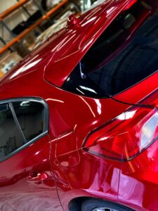 Close-up of the rear section of a shiny red car, showcasing the sleek, reflective surface and modern design elements. The background includes a storage area with shelves and various items, indicating an indoor garage or workshop setting.