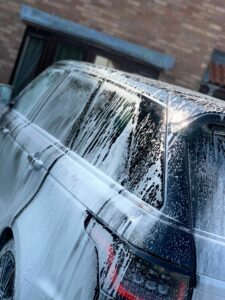 A black SUV covered in soap suds is parked outside, adjacent to a brick building. It appears to be in the process of being washed. Sunlight reflects off the wet surface of the vehicle, highlighting the suds and soap streaks.