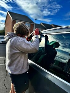 A person in a gray hoodie and red gloves is working on a car window with a tool. The person is partially bent over and facing the vehicle. The background shows a suburban setting with a house and a cloudy sky.