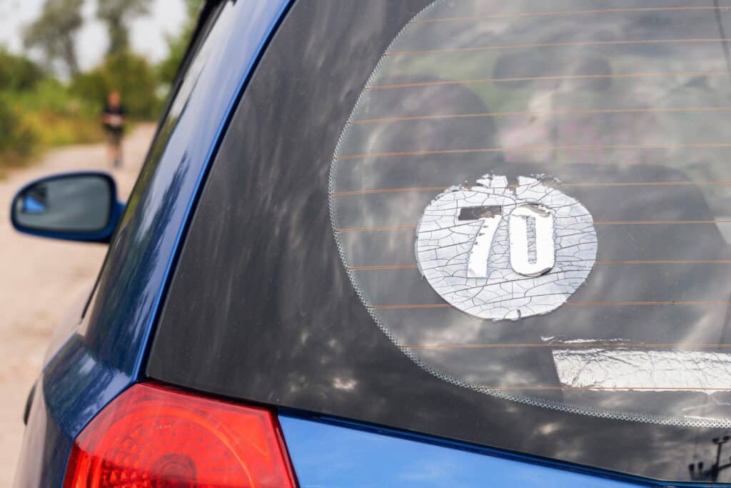 A close-up of a blue car's rear window reveals a cracked sticker with the number "70." The circular, white decal hints at the need for cleaning services. Blurred greenery and a person walking enhance the scene in the background.